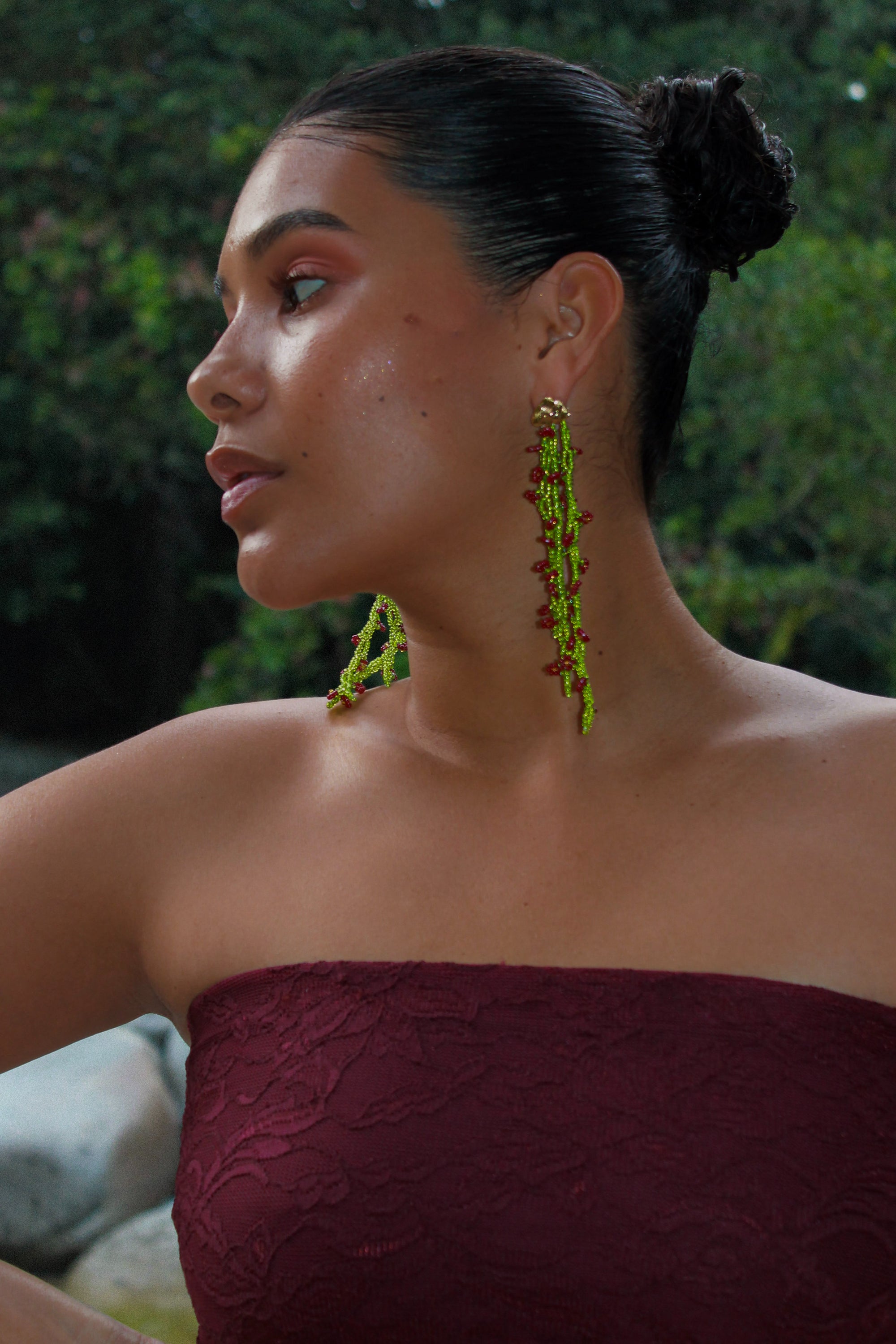 Woman wearing a strapless maroon top with green earrings against a blurred natural background