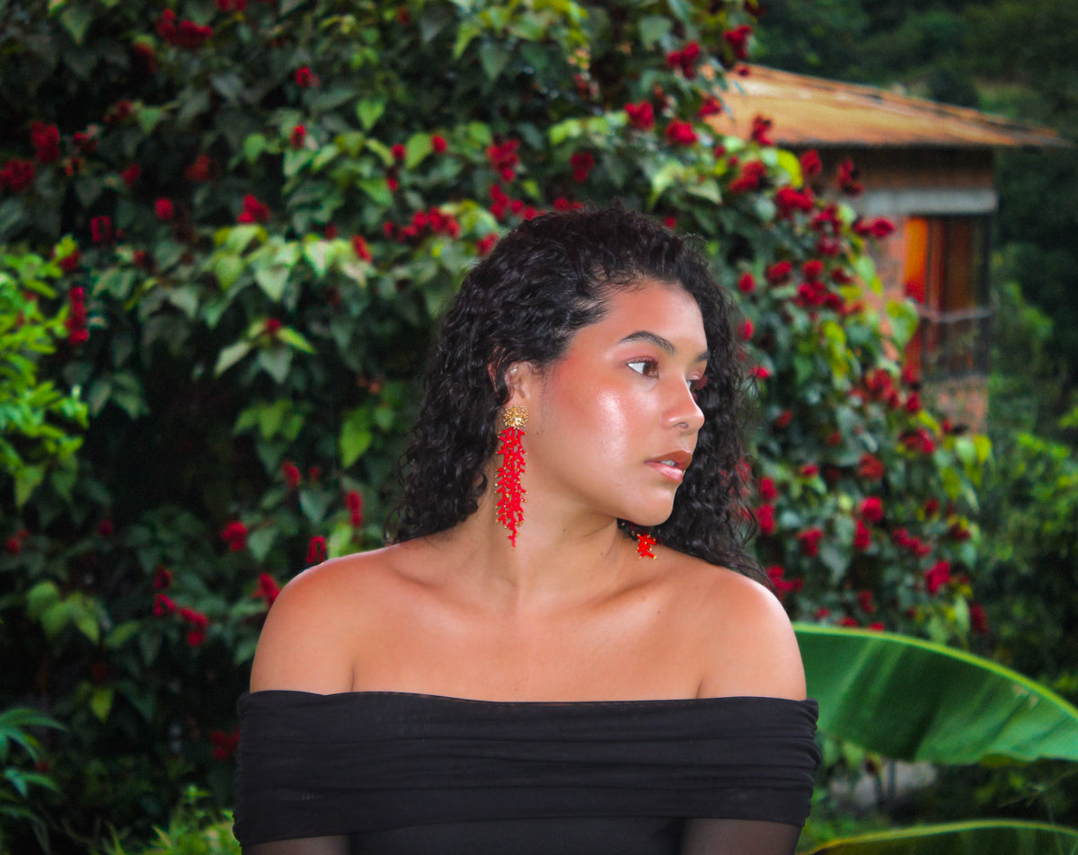 Woman wearing a black off-shoulder top with red earrings in front of green foliage with red flowers.