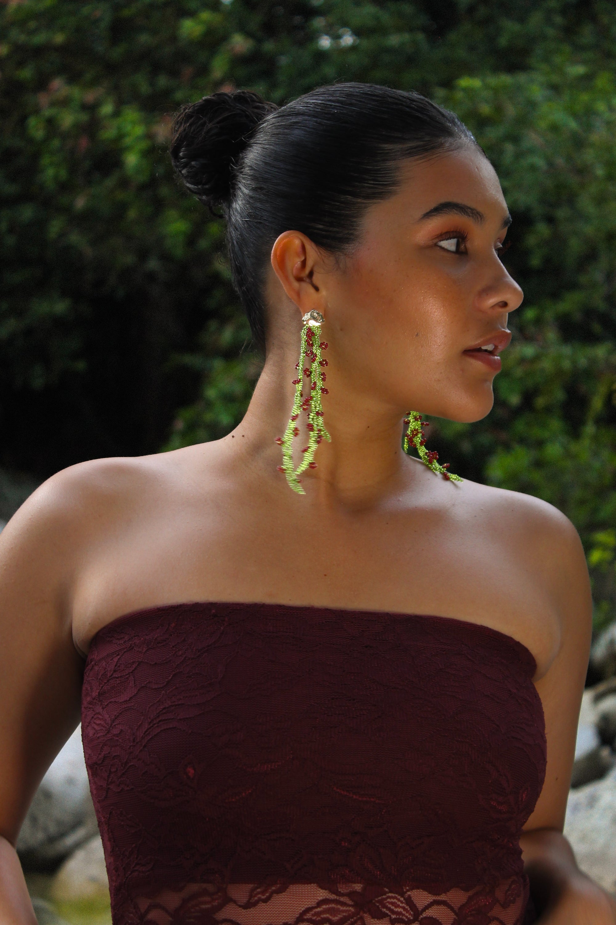 Woman wearing a strapless burgundy dress with green earrings against a natural background