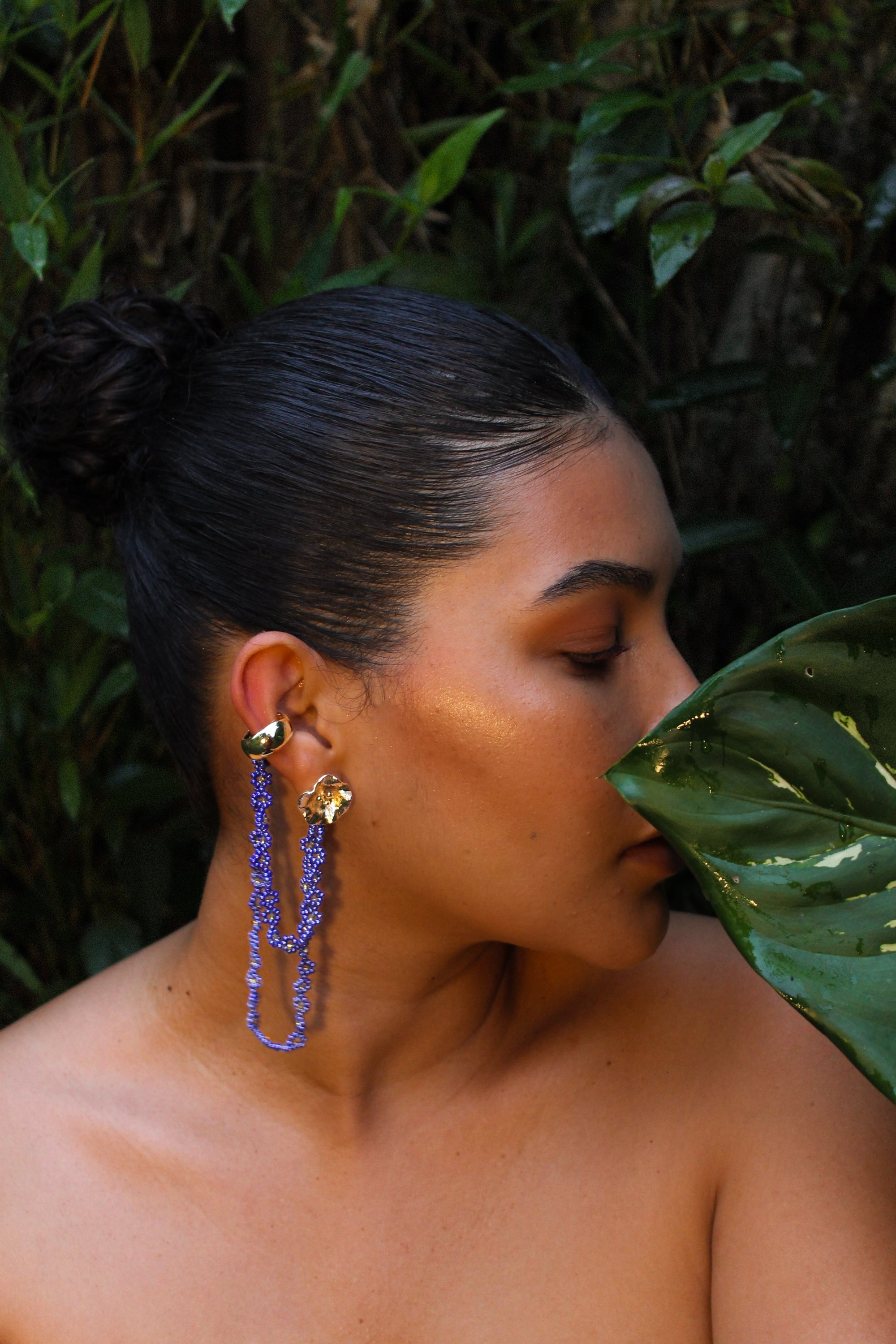 Woman wearing long blue earrings with a green leaf in front of her face, against a natural background.
