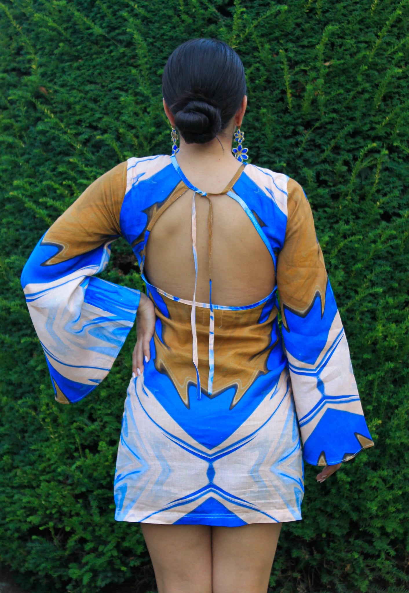 Person wearing a blue and brown dress with a geometric pattern against a green hedge.